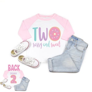 May include: A white and pink long-sleeved shirt with a donut graphic and the text "TWO sassy and sweet". The shirt is a baseball style with pink sleeves and a white body. The shirt is laid out on a white background with a pair of white sneakers and blue jeans.