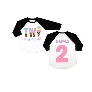 Pu&ograve; includere: Due magliette da baseball a tema cono gelato. La parte anteriore di una maglietta dice "Two sassy and sweet" con una grafica di cono gelato. La parte posteriore dell'altra maglietta dice "Emma 2" in rosa.
