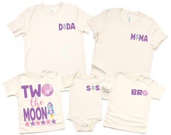 Puede incluir: Cinco camisetas color crema y un body con texto y gráficos morados. Las camisetas dicen "DADA", "MAMA", "TWO the MOON" y "BRO". El body dice "S/S". Todos tienen un gráfico de cohete.