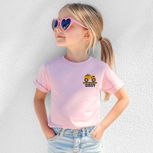 May include: A light pink short-sleeve t-shirt featuring a small graphic on the left chest. The graphic displays a yellow dump truck above black and yellow caution stripes and the word SISSY printed in bold black capital letters.