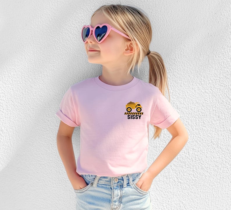 May include: A light pink short-sleeve t-shirt featuring a small graphic on the left chest. The graphic displays a yellow dump truck above black and yellow caution stripes and the word SISSY printed in bold black capital letters.