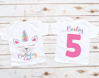 May include: Two white t-shirts, one with a unicorn cat design and the text "Birthday Girl" and the other with the name "Bexley" and the number "5" in pink.