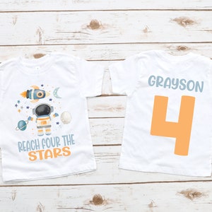 May include: White t-shirt with a blue and orange astronaut design and the text "Reach four the stars". The back of the shirt has the name "Grayson" and the number "4" in orange.