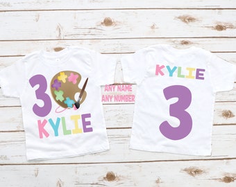 Può includere: Due magliette bianche con un numero 3 viola e il nome Kylie in colori arcobaleno. La maglietta anteriore ha una tavolozza di colori marrone con schizzi di vernice colorati e un pennello.