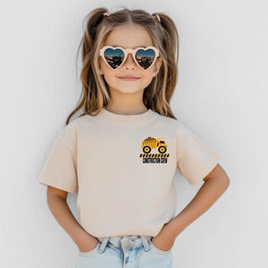 May include: A beige short-sleeved t-shirt featuring a small graphic on the left chest. The graphic depicts a yellow dump truck carrying brown dirt, positioned above black and yellow hazard stripes and the text CONSTRUCTION CREW in black.