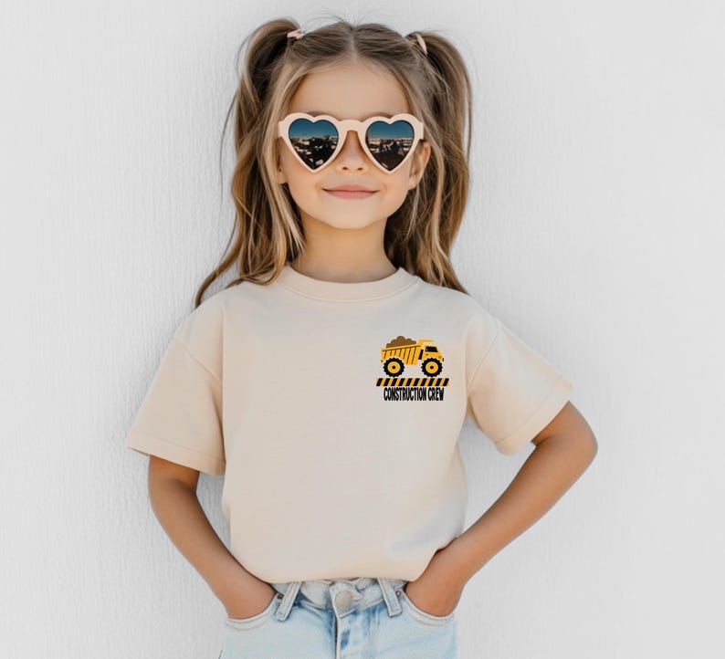 May include: A beige short-sleeved t-shirt featuring a small graphic on the left chest. The graphic depicts a yellow dump truck carrying brown dirt, positioned above black and yellow hazard stripes and the text CONSTRUCTION CREW in black.