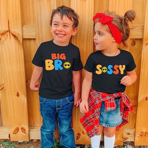 May include: Two black t-shirts featuring colourful graphic text. The left shirt displays the text BIG BRO with a sun icon wearing sunglasses. The right shirt displays the text SASSY with a sun icon wearing sunglasses in place of the letter A.
