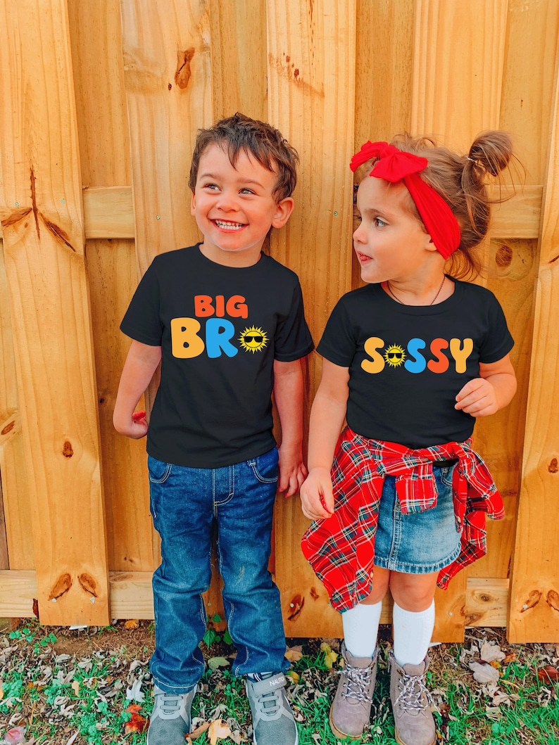 May include: Two black t-shirts featuring colourful graphic text. The left shirt displays the text BIG BRO with a sun icon wearing sunglasses. The right shirt displays the text SASSY with a sun icon wearing sunglasses in place of the letter A.