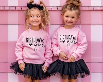 May include: Two young girls wearing matching pink sweatshirts with the text "BIRTHDAY girl" in black script and a heart. They are also wearing black tulle skirts. The background is a pink and white striped wall.
