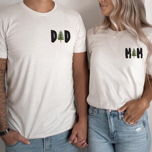 Puede incluir: Dos camisetas crema a juego. Una camiseta tiene la palabra "DAD" con un gr&aacute;fico de &aacute;rbol verde, la otra dice "MOM" con un &aacute;rbol similar. Ambas camisetas son de manga corta y se usan con jeans azules.