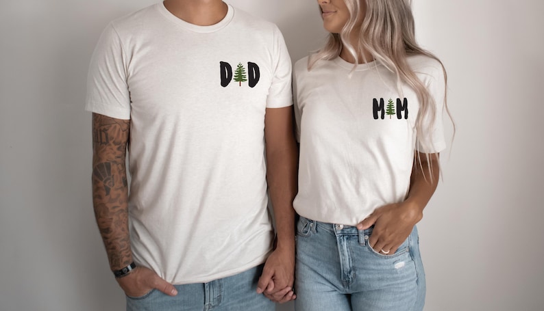 Puede incluir: Dos camisetas crema a juego. Una camiseta tiene la palabra "DAD" con un gr&aacute;fico de &aacute;rbol verde, la otra dice "MOM" con un &aacute;rbol similar. Ambas camisetas son de manga corta y se usan con jeans azules.