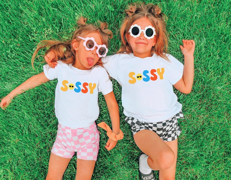 May include: Two white short-sleeved t-shirts featuring the word SASSY printed in colourful, rounded letters. The letter A is replaced by a yellow smiley face wearing black sunglasses. The shirts are paired with pink and white chequered shorts.