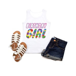 May include: White tank top with the text "Birthday Girl" in pink and colourful block letters. The letters are made up of small colourful blocks. The tank top is lying on a white surface with a pair of white sandals and blue denim shorts.