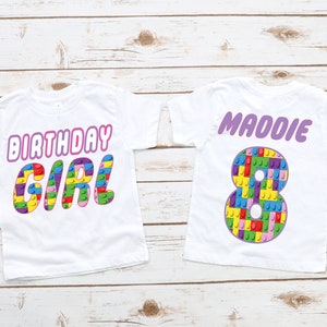 May include: Two white t-shirts with a birthday theme. The first shirt says "Birthday Girl" in pink letters with a colourful Lego block design. The second shirt has a large number 8 made of Lego blocks and the name "Maddie" in purple letters.