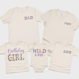 May include: A collection of five cream-colored garments including two adult t-shirts with floral DAD and MOM text, a child's t-shirt with purple and floral Birthday GIRL text, a baby bodysuit with purple and floral WILD one text, and a child's t-shirt with floral BRO text.