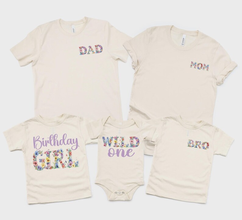May include: A collection of five cream-colored garments including two adult t-shirts with floral DAD and MOM text, a child's t-shirt with purple and floral Birthday GIRL text, a baby bodysuit with purple and floral WILD one text, and a child's t-shirt with floral BRO text.
