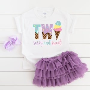 Pu&ograve; includere: Maglietta bianca con il testo "TWO" in lettere viola e blu con coni gelato. Il testo "sassy and sweet" &egrave; sotto il numero. La maglietta &egrave; abbinata a una gonna di tulle viola.