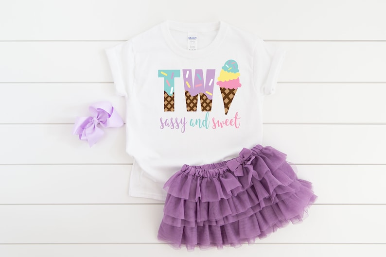 Pu&ograve; includere: Maglietta bianca con il testo "TWO" in lettere viola e blu con coni gelato. Il testo "sassy and sweet" &egrave; sotto il numero. La maglietta &egrave; abbinata a una gonna di tulle viola.