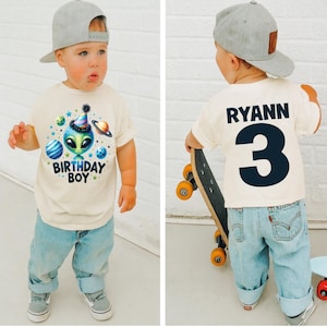 May include: A toddler wearing a cream-colored t-shirt with an alien graphic and the text "BIRTHDAY BOY." The back of the shirt has the name "RYANN" and the number "3." The child is wearing light blue jeans and a gray baseball cap.