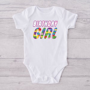 May include: White baby onesie with the text "Birthday Girl" in pink letters. The letters are made up of colourful Lego blocks.