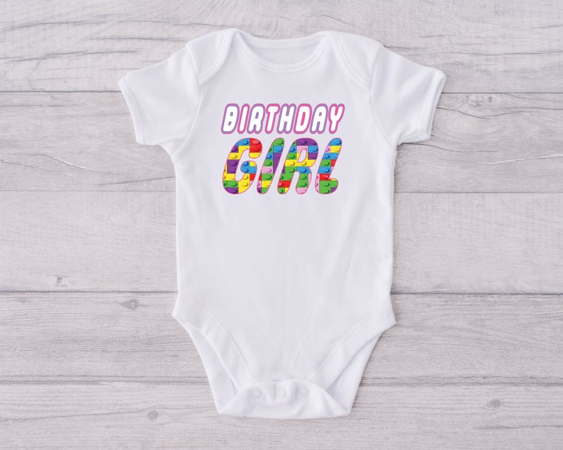 May include: White baby onesie with the text "Birthday Girl" in pink letters. The letters are made up of colourful Lego blocks.