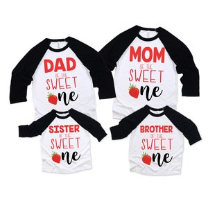 K&ouml;nnte beinhalten: Set aus vier wei&szlig;en T-Shirts mit schwarzen &Auml;rmeln. Die Shirts haben roten Text, der "Papa des s&uuml;&szlig;en Kleinen", "Mama des s&uuml;&szlig;en Kleinen", "Schwester des s&uuml;&szlig;en Kleinen" und "Bruder des s&uuml;&szlig;en Kleinen" lautet. Jedes Shirt hat eine rote Erdbeergrafik.