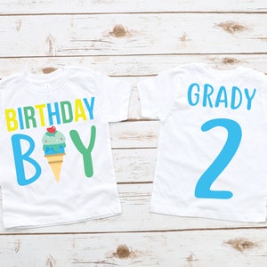 May include: Two white t-shirts with blue, green and yellow text and an ice cream cone graphic. The first shirt says "Birthday Boy" and the second shirt says "Grady 2".