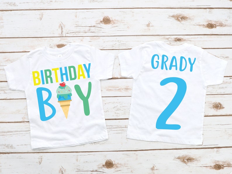 May include: Two white t-shirts with blue, green and yellow text and an ice cream cone graphic. The first shirt says "Birthday Boy" and the second shirt says "Grady 2".