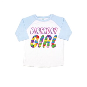 May include: A white and light blue raglan t-shirt with the words "Birthday Girl" in pink letters. The letters are made up of colourful Lego blocks.
