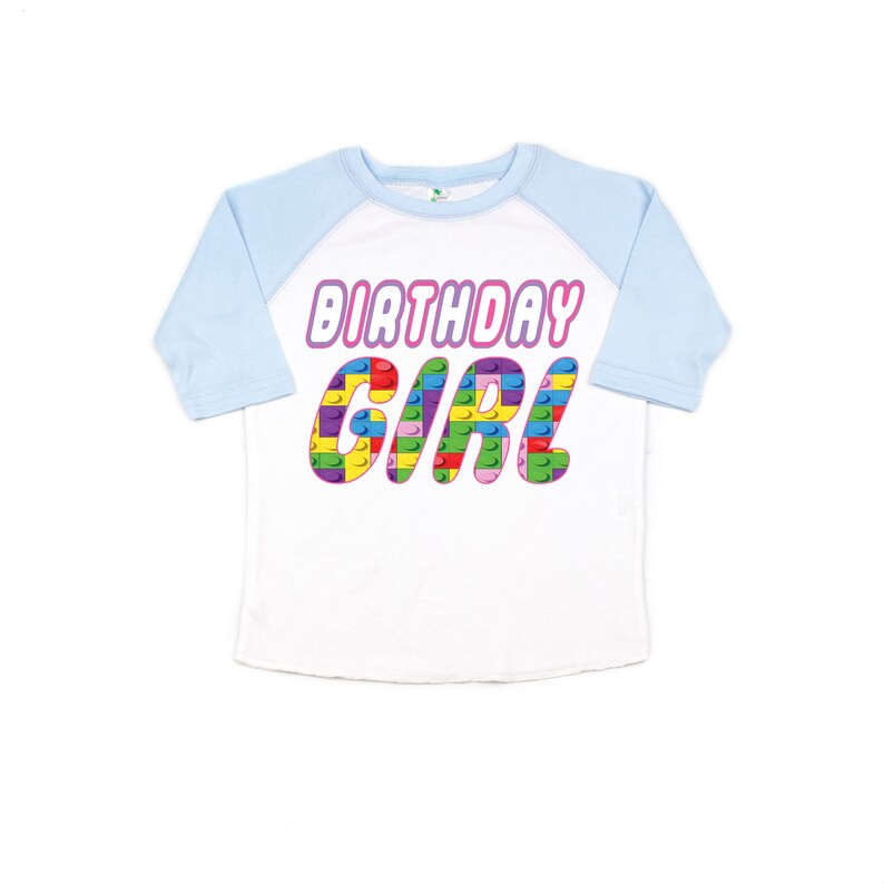 May include: A white and light blue raglan t-shirt with the words "Birthday Girl" in pink letters. The letters are made up of colourful Lego blocks.