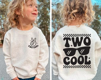 Puede incluir: Una sudadera de color beige claro con un gráfico negro en la parte delantera que dice "Two Cool" con un signo de paz. La parte posterior de la sudadera tiene un gráfico negro que dice "TWO COOL" con gafas de sol y un patrón a cuadros.