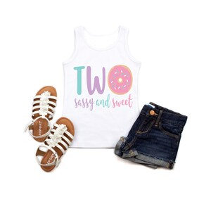 May include: White tank top with the text "TWO sassy and sweet" and a pink donut with sprinkles. The tank top is paired with denim shorts and white sandals.