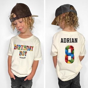 May include: A white t-shirt with the words "Birthday Boy" in colorful block letters. The name "Adrian" is printed below the text. The back of the shirt has the name "Adrian" printed in black and the number "8" made of colorful blocks.