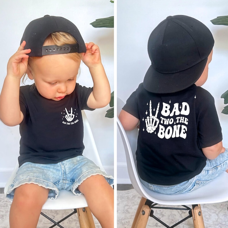 Puede incluir: Una camiseta negra con texto blanco que dice "Bad Two The Bone" y una mano de esqueleto haciendo un signo de paz. La camiseta la lleva un ni&ntilde;o que lleva una gorra de b&eacute;isbol negra.