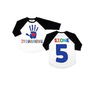 May include: A white and black baseball style shirt with a blue handprint and the text "I'm a whole handful" on the front. The back of the shirt has the name "SIONE" and the number "5" in blue.
