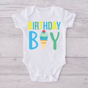 May include: White baby onesie with the text "BIRTHDAY BOY" in yellow, blue and green letters. An ice cream cone with a cherry on top is between the "B" and the "Y".