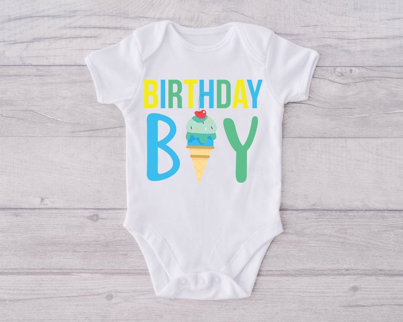 May include: White baby onesie with the text "BIRTHDAY BOY" in yellow, blue and green letters. An ice cream cone with a cherry on top is between the "B" and the "Y".