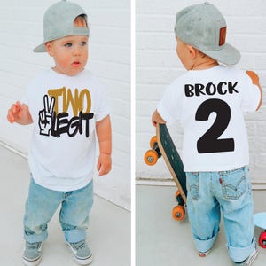 Pu&ograve; includere: Un bambino piccolo indossa una maglietta bianca con la scritta "TWO LEGIT" in nero e oro, abbinata a jeans azzurri e un berretto da baseball grigio. Il bambino indossa anche scarpe da ginnastica grigie. La parte posteriore della maglietta ha il nome "BROCK" e il numero "2".