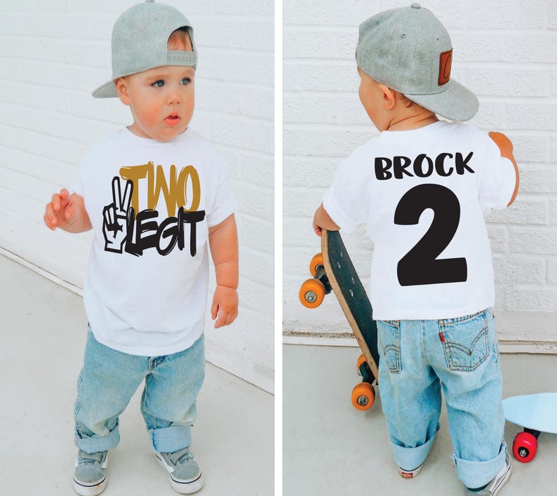 Pu&ograve; includere: Un bambino piccolo indossa una maglietta bianca con la scritta "TWO LEGIT" in nero e oro, abbinata a jeans azzurri e un berretto da baseball grigio. Il bambino indossa anche scarpe da ginnastica grigie. La parte posteriore della maglietta ha il nome "BROCK" e il numero "2".
