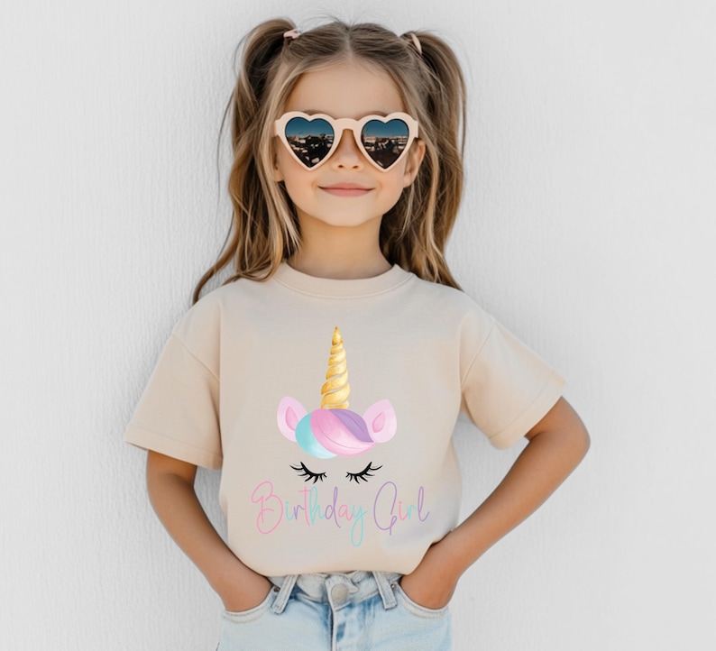 May include: A young girl wearing heart-shaped sunglasses and a beige t-shirt. The t-shirt features a unicorn graphic with a golden horn, pastel ears, and the text "Birthday Girl" in a script font.