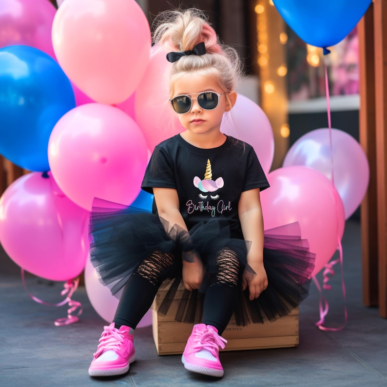 May include: A young person wearing sunglasses, a black t-shirt with a unicorn graphic, a black tutu, and ripped black leggings. The person is wearing pink sneakers and is surrounded by pink and blue balloons. The text on the shirt says "Birthday Girl."