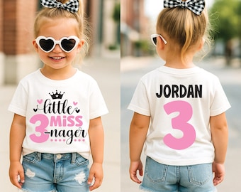 May include: A young child wearing a white t-shirt with the text "little miss 3-nager" in pink and black. The child is also wearing denim shorts, heart-shaped sunglasses, and a black and white checkered bow.