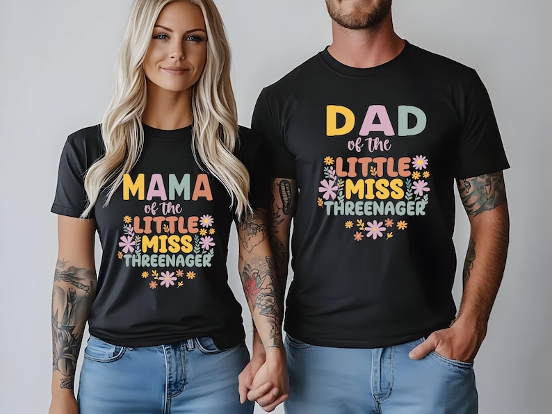 May include: Two black short-sleeve t-shirts featuring colorful floral graphics and text. One shirt displays 'MAMA of the LITTLE MISS THREENAGER' and the other displays 'DAD of the LITTLE MISS THREENAGER' in a mix of pastel and bold lettering.