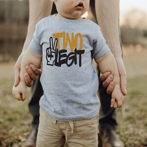 Pu&ograve; includere: Una maglietta grigia a maniche corte con la scritta "TWO LEGIT" in nero e giallo. La maglietta presenta una grafica con il segno della pace. Il bambino indossa pantaloncini color cachi. Lo sfondo &egrave; sfocato, con una persona che tiene le mani del bambino.
