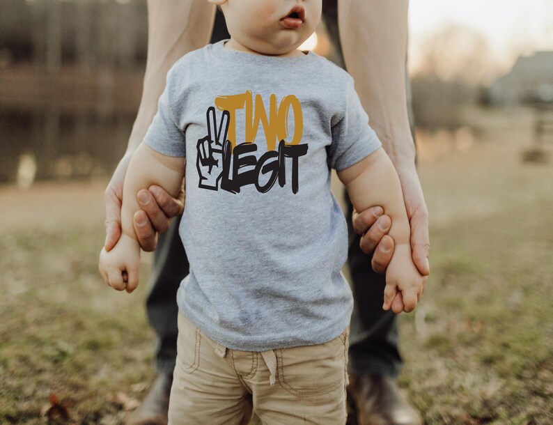 Pu&ograve; includere: Una maglietta grigia a maniche corte con la scritta "TWO LEGIT" in nero e giallo. La maglietta presenta una grafica con il segno della pace. Il bambino indossa pantaloncini color cachi. Lo sfondo &egrave; sfocato, con una persona che tiene le mani del bambino.
