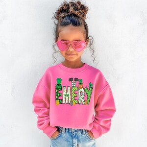 May include: A pink sweatshirt with a green, yellow, and orange graphic of the name "EMERY" with leprechauns and shamrocks. The sweatshirt is being worn by a young girl with curly brown hair and pink heart-shaped sunglasses.