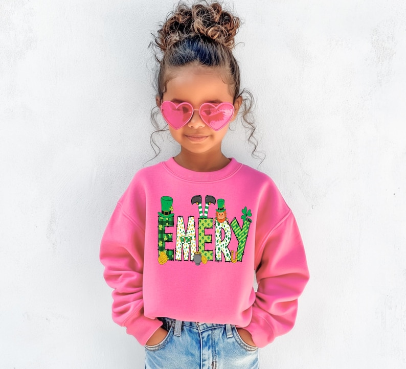 May include: A pink sweatshirt with a green, yellow, and orange graphic of the name "EMERY" with leprechauns and shamrocks. The sweatshirt is being worn by a young girl with curly brown hair and pink heart-shaped sunglasses.