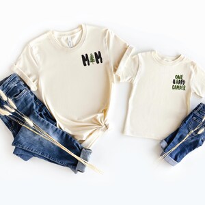 Puede incluir: Dos camisetas color crema con los textos "MOM" y "ONE HAPPY CAMPER" en negro y verde, junto con un peque&ntilde;o gr&aacute;fico de &aacute;rbol. Las camisetas se combinan con vaqueros azules. Una ramita de flores secas descansa sobre los vaqueros.