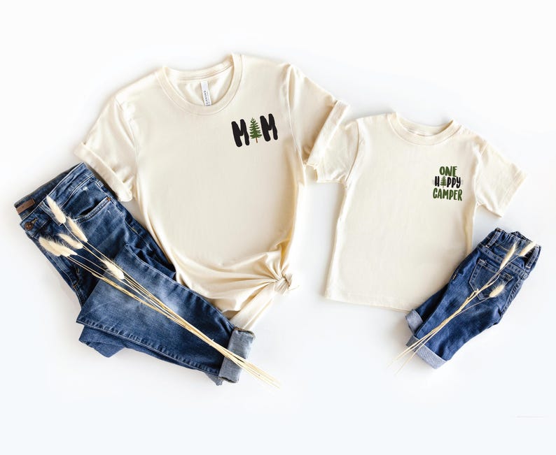 Puede incluir: Dos camisetas color crema con los textos "MOM" y "ONE HAPPY CAMPER" en negro y verde, junto con un peque&ntilde;o gr&aacute;fico de &aacute;rbol. Las camisetas se combinan con vaqueros azules. Una ramita de flores secas descansa sobre los vaqueros.