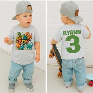 May include: A light gray t-shirt with a colorful dinosaur graphic and the text "BIRTHDAY BOY." The back of the shirt has the name "RYANN" and the number "3" in green. The child is wearing light blue jeans and a gray baseball cap.